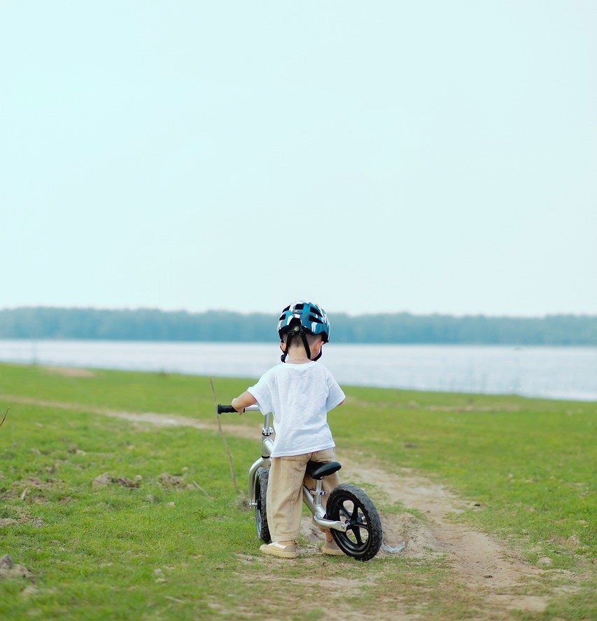 Ein kleines Kind in einem weißen Shirt und einem Fahrradhelm steht auf einem Pfad neben einem Fluss und hält einen Stock in der Hand.